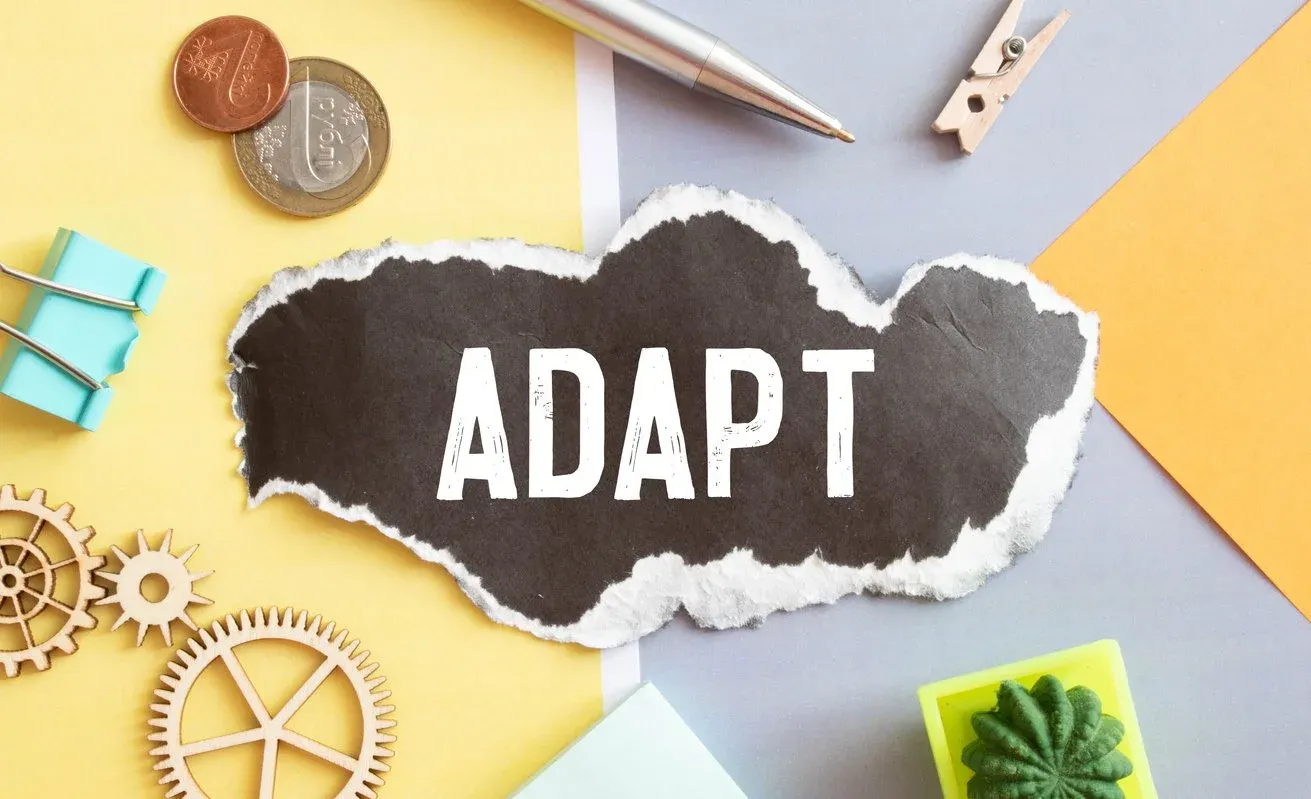 The word "Adapt" on a torn scrap of black paper, surrounded by wooden gears, coins, and desk supplies.